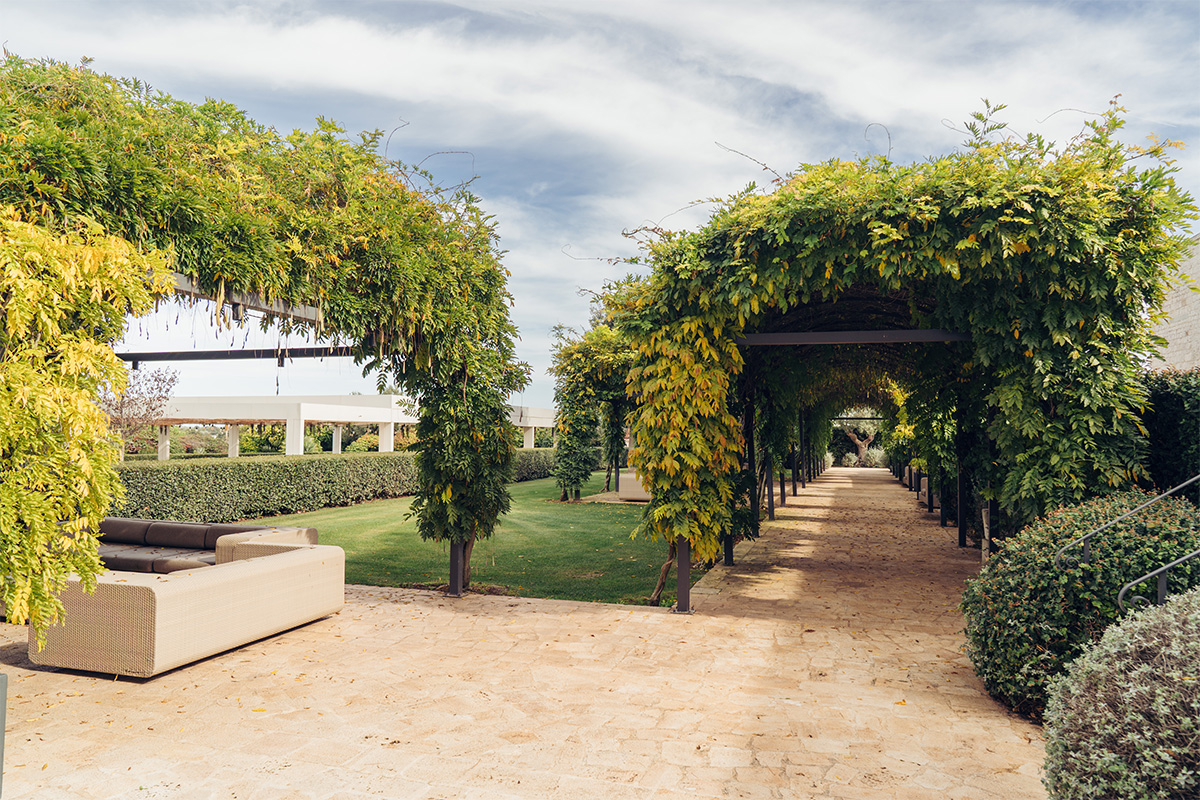 Pergolato verde nel giardino, con vialetto in pietra, piante fiorite e area lounge all’aperto.