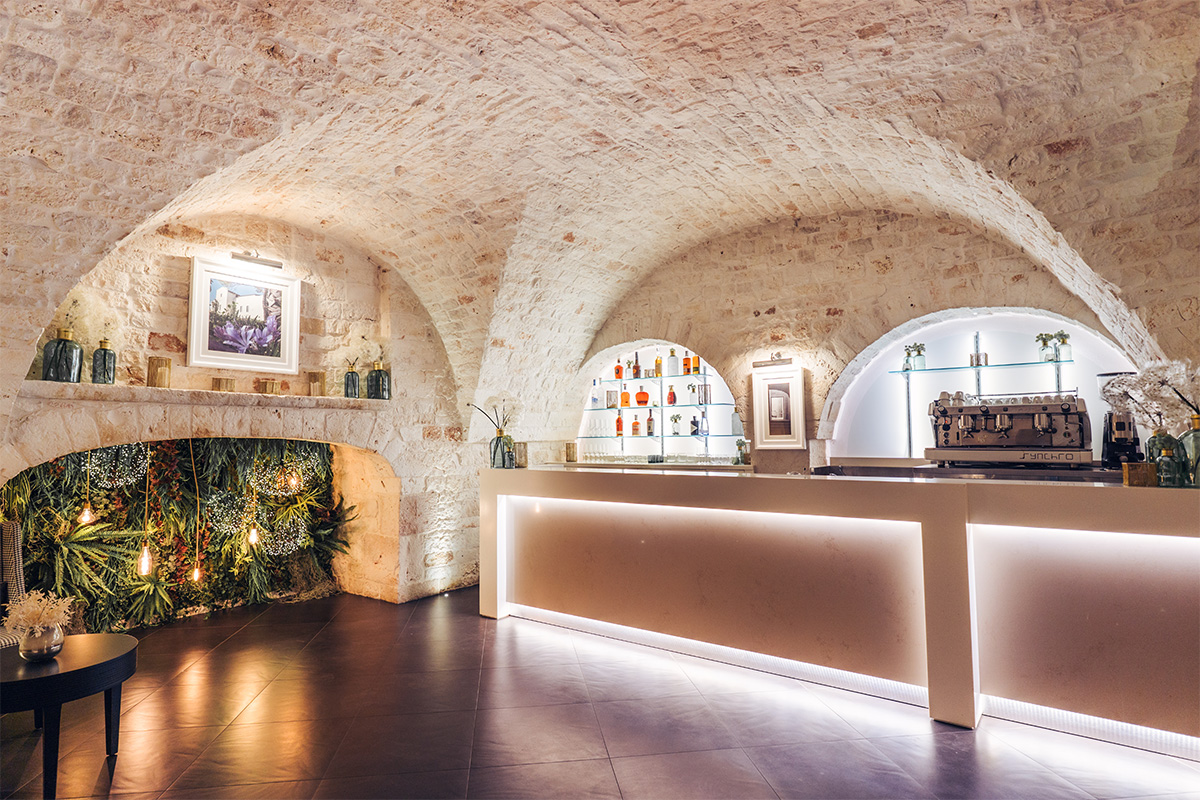 Area bar dell’hotel Il Santissimo con bancone illuminato, volte in pietra e dettagli storici.