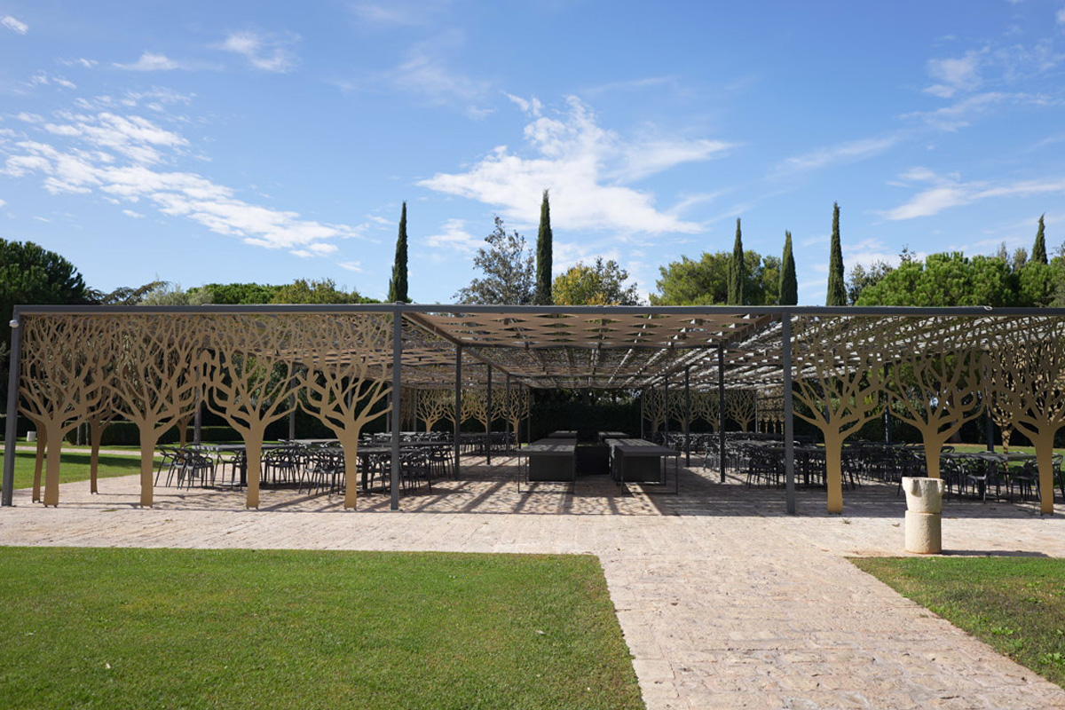Shaded pergola in the park, with outdoor seating, lawn, stone path and cypress trees in the background.